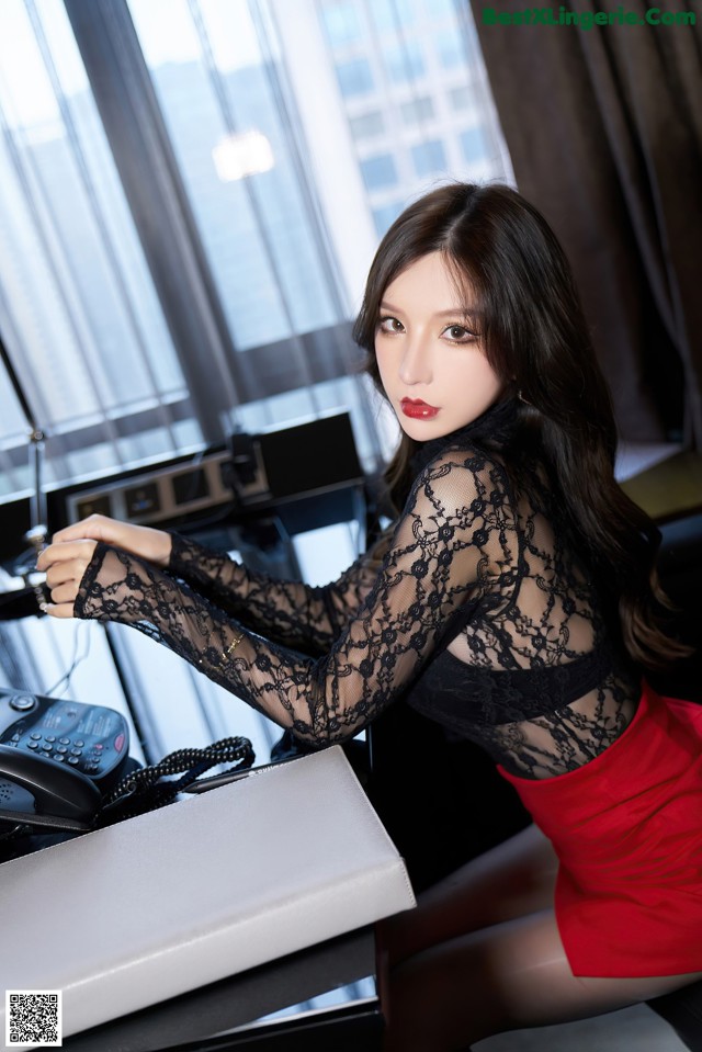 A woman in a black lace top and red skirt sitting at a desk.