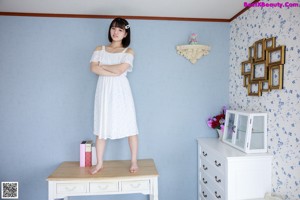 A woman in a white dress standing next to a dresser.