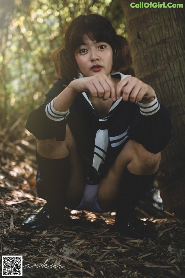 A woman in a school uniform crouching down in the woods.