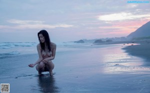 A naked woman sitting on a beach next to the ocean.